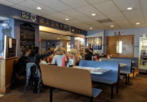 A cozy diner interior with several tables, patrons chatting, and a server attending to customers. Walls adorned with plaques.