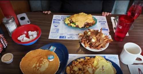 A table set with a large plate of cheesy breakfast, pancakes, fries, and a cup of coffee, with condiments nearby.