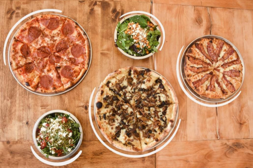 A wooden table with four pizzas and two bowls of salad, showcasing various toppings and styles.