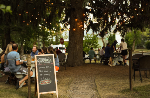Outdoor dining scene with people at tables under a tree, string lights, and a sign welcoming guests to Sidewall.