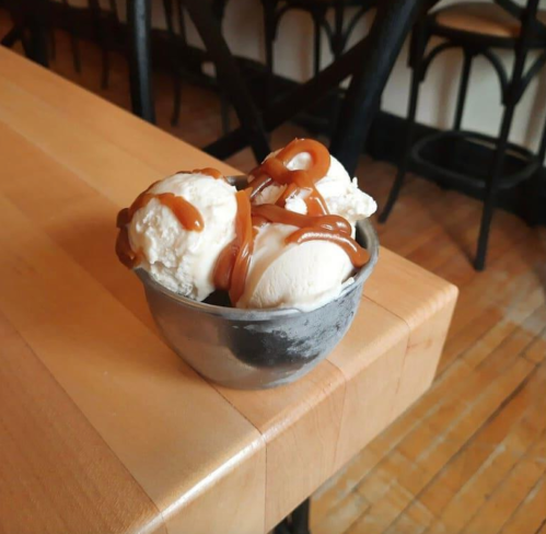 A small metal bowl filled with three scoops of vanilla ice cream drizzled with caramel sauce, placed on a wooden table.