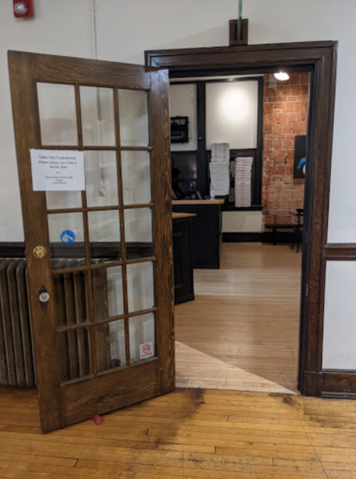A wooden door with glass panels opens to a room with a brick wall and a desk, featuring a sign on the door.