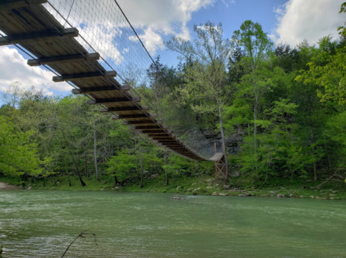 A wooden suspension bridge spans a green river, surrounded by lush trees and a blue sky with scattered clouds.