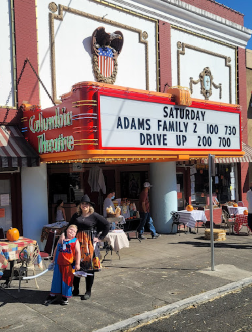 A woman and child in costumes stand outside the Columbia Theatre, with a marquee displaying "Adams Family 2" showtimes.