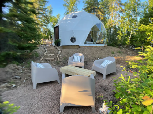 A geodesic dome home surrounded by trees, with four outdoor chairs and a wooden table in a natural setting.