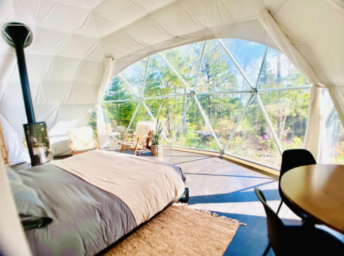 Cozy interior of a geodesic dome with large windows, a bed, and a small dining area surrounded by nature.