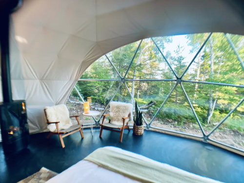 Cozy interior of a geodesic dome with two chairs, a small table, and large windows overlooking a forested area.
