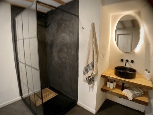 Modern bathroom featuring a glass shower, round mirror, and wooden shelf with toiletries. Neutral color scheme.