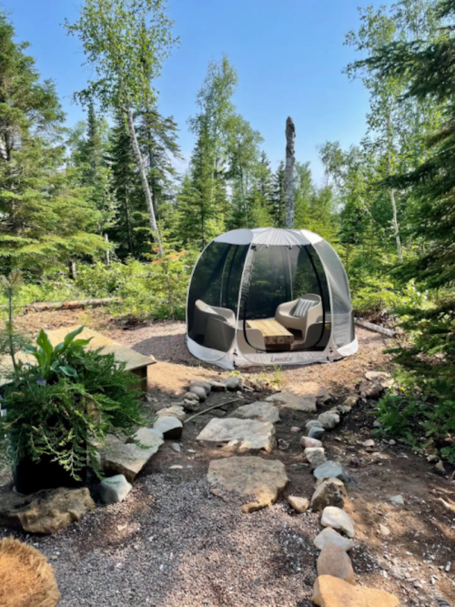 A cozy outdoor seating area in a forest, featuring a transparent tent and stone pathway surrounded by greenery.