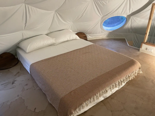 A cozy bedroom with a large bed, white bedding, and a round window in a dome-shaped room.