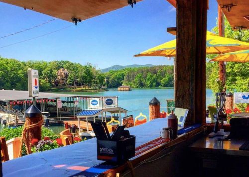 View from a restaurant overlooking a serene lake, with boats and lush greenery in the background. Bright umbrellas add color.