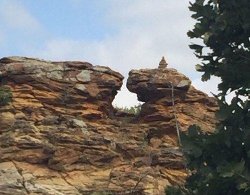 A rocky outcrop with a small stone stack on top, surrounded by greenery and a blue sky.