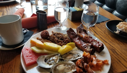 A plate of assorted food including ribs, oysters, fruit, and a meatball, with drinks and table settings in the background.