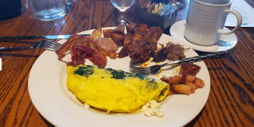 A plate with a spinach omelet, bacon, sausage, potatoes, and fruit, accompanied by a cup and utensils on a wooden table.