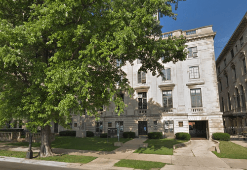 A large, historic building with a stone facade, surrounded by green trees and a clear blue sky.