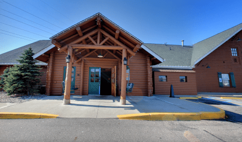 A log cabin-style building with a large entrance and wooden pillars, surrounded by a paved area and greenery.