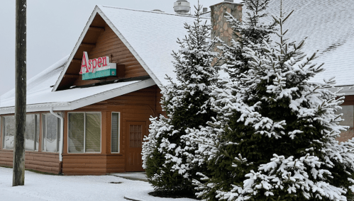 A snow-covered building with a sign reading "Aspen" and evergreen trees in front.