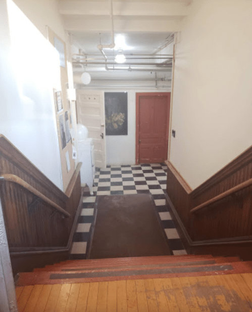 A staircase leading down to a checkered floor hallway with doors and a light fixture above.