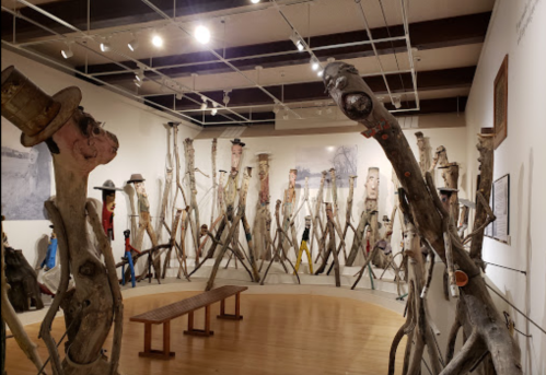 A gallery filled with wooden sculptures resembling figures, some wearing hats, arranged in a circular layout.