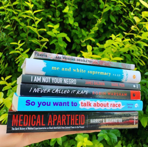 A stack of books on race and social justice against a green leafy background. Titles include "Medical Apartheid" and "I Am Not Your Negro."
