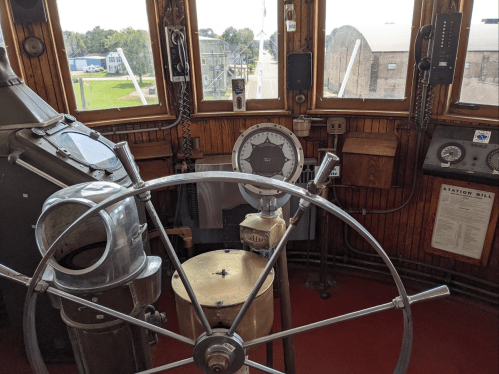 A vintage ship's wheel and control panel inside a cabin, with gauges and a view of the outdoors through windows.