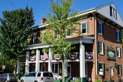 Historic brick building with green shutters, white columns, and patriotic decorations, surrounded by trees and parked cars.