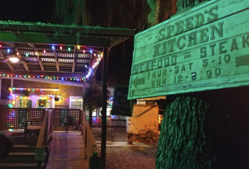 A brightly lit outdoor restaurant sign for "Speed's Kitchen," featuring seafood and steak, with festive lights in the background.