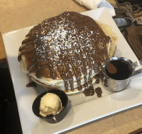 A stack of pancakes drizzled with chocolate sauce, topped with powdered sugar, served with a side of butter and syrup.
