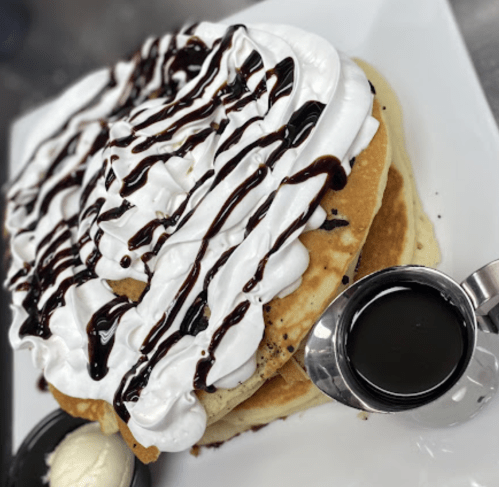 A stack of fluffy pancakes topped with whipped cream, chocolate drizzle, and a side of syrup and ice cream.