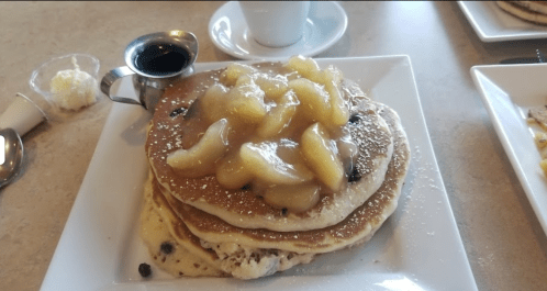Stack of pancakes topped with apple slices and syrup, served on a white plate with a cup in the background.