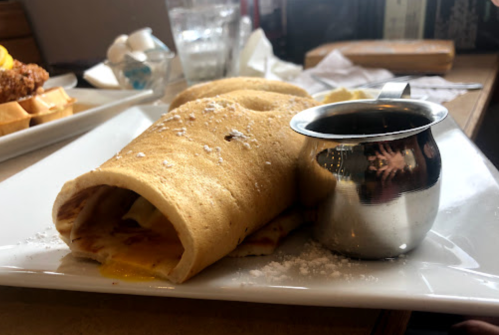 A rolled crepe filled with eggs, served on a plate with a small metal jug of syrup.
