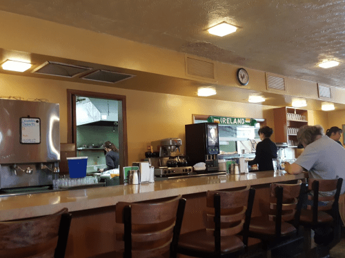 A cozy diner interior with a counter, coffee machine, and patrons seated, while staff prepare food in the kitchen.