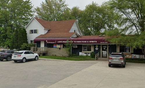 A roadside tavern with a red awning, surrounded by trees and parked cars in a gravel lot.
