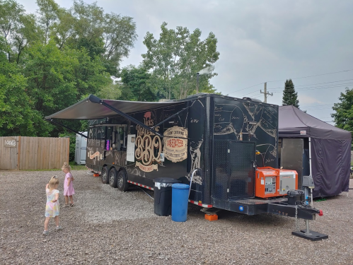 A food truck with a black exterior parked in a gravel area, with two children playing nearby under a cloudy sky.