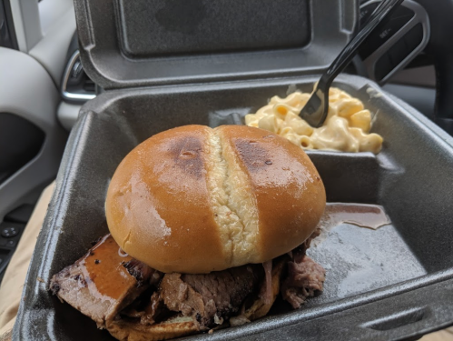 A takeout container with a sandwich filled with meat and a side of macaroni and cheese.
