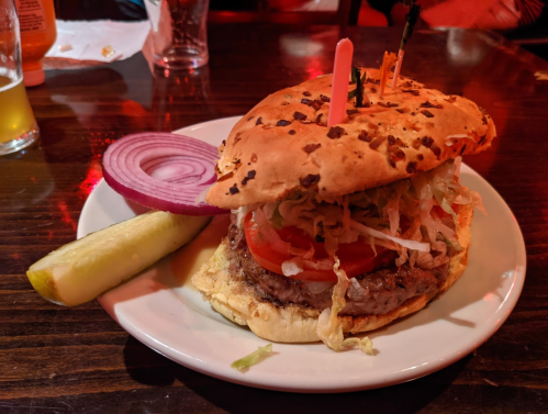 A large burger with lettuce, tomato, and onion on a plate, accompanied by a pickle and a slice of red onion.