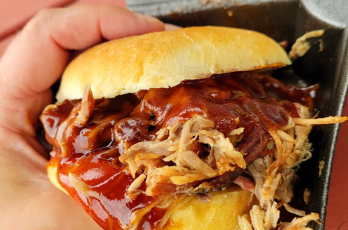 A hand holding a pulled pork sandwich topped with barbecue sauce on a soft bun.