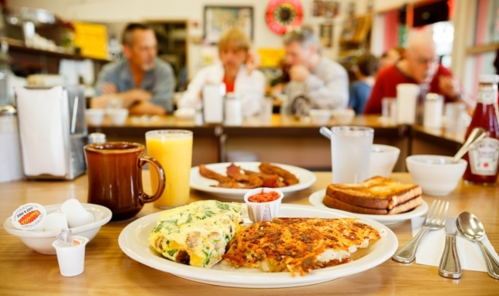 A breakfast spread featuring an omelet, hash browns, toast, and drinks, with diners in the background.