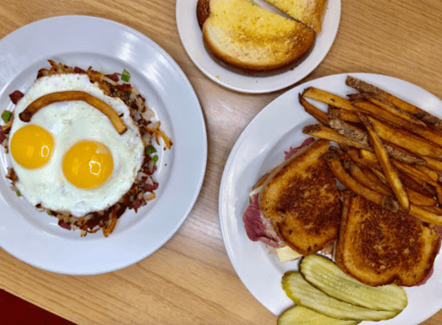 A plate with two sunny-side-up eggs on hash browns, and another plate with grilled cheese, fries, and pickles.