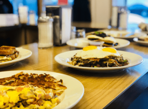 A table with various breakfast dishes, including eggs, hash browns, and sandwiches, in a cozy diner setting.