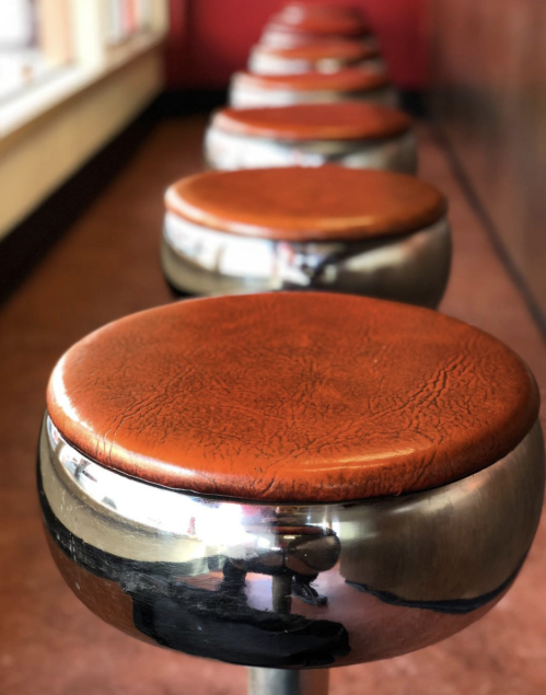 A row of shiny metal stools with brown leather seats, lined up against a red wall.