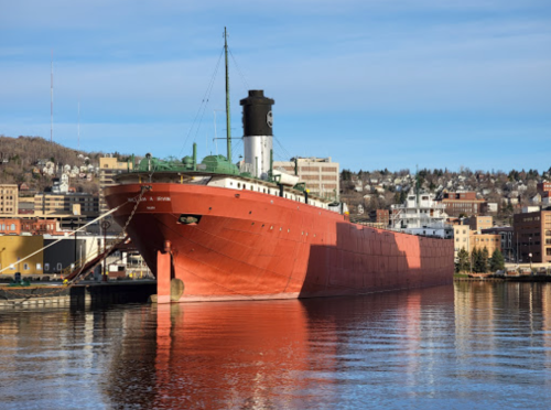 A large red ship docked in a harbor with buildings and hills in the background under a clear blue sky.