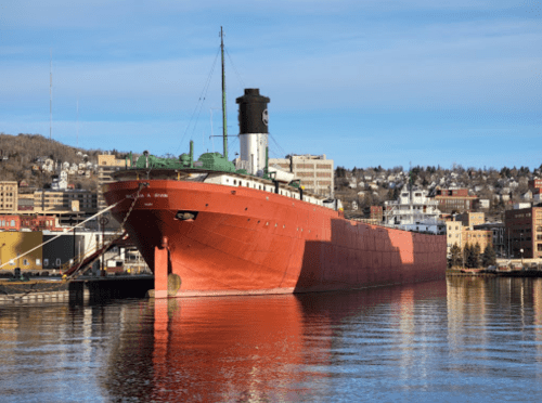 A large red ship docked in a harbor with buildings and hills in the background under a clear blue sky.