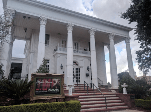 A large white mansion with columns, featuring a staircase and a sign for a live event. Lush greenery surrounds the building.