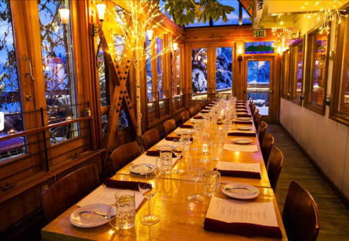 A warmly lit restaurant with a long wooden table set for dining, surrounded by windows and festive lights.