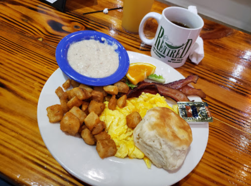 A plate of breakfast featuring scrambled eggs, bacon, potatoes, a biscuit, grits, and an orange slice, with coffee nearby.