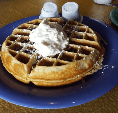 A golden waffle topped with whipped cream and powdered sugar, served on a blue plate with syrup containers in the background.