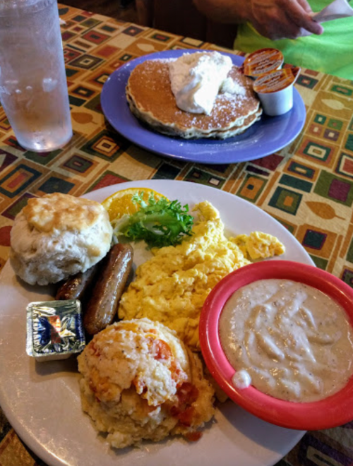 A plate of breakfast featuring biscuits, sausage, scrambled eggs, grits, and a side of pancakes with whipped cream.