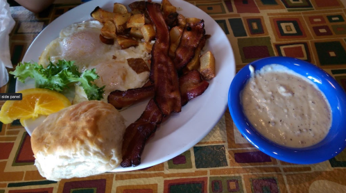 A plate of breakfast featuring eggs, bacon, potatoes, a biscuit, and a bowl of gravy, with a slice of orange and lettuce.