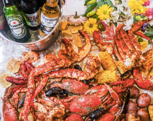 A vibrant seafood spread featuring crabs, shrimp, corn, potatoes, and drinks on a newspaper backdrop.